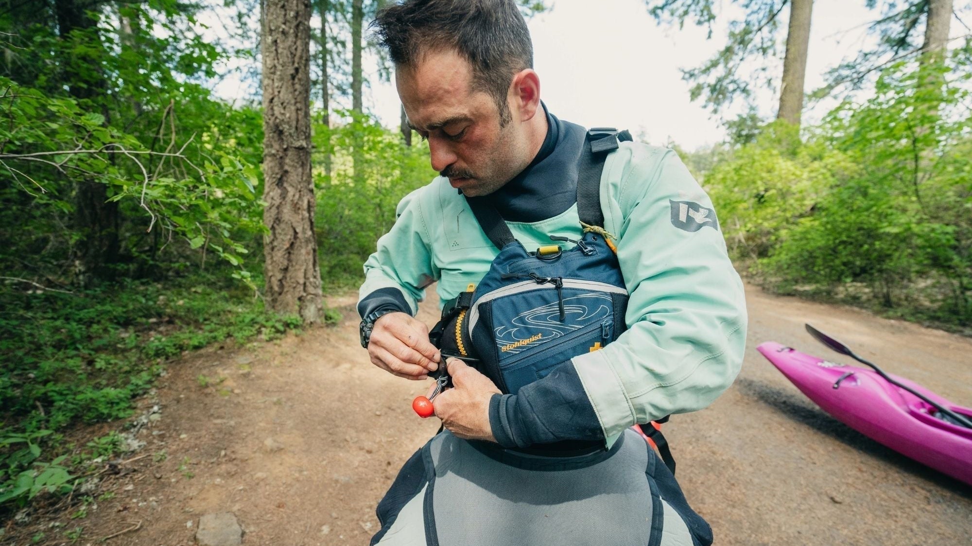 Man wearing Stohlquist kayak life jacket adjusting gear outdoors beside pink kayak