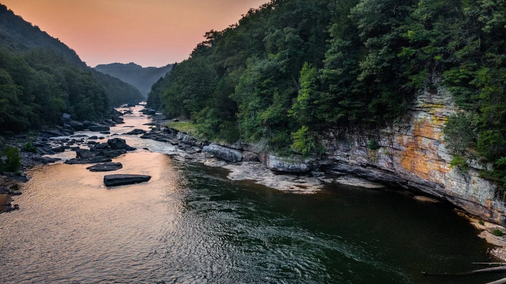 River flowing through forested rocky canyon at sunset with orange sky and green trees