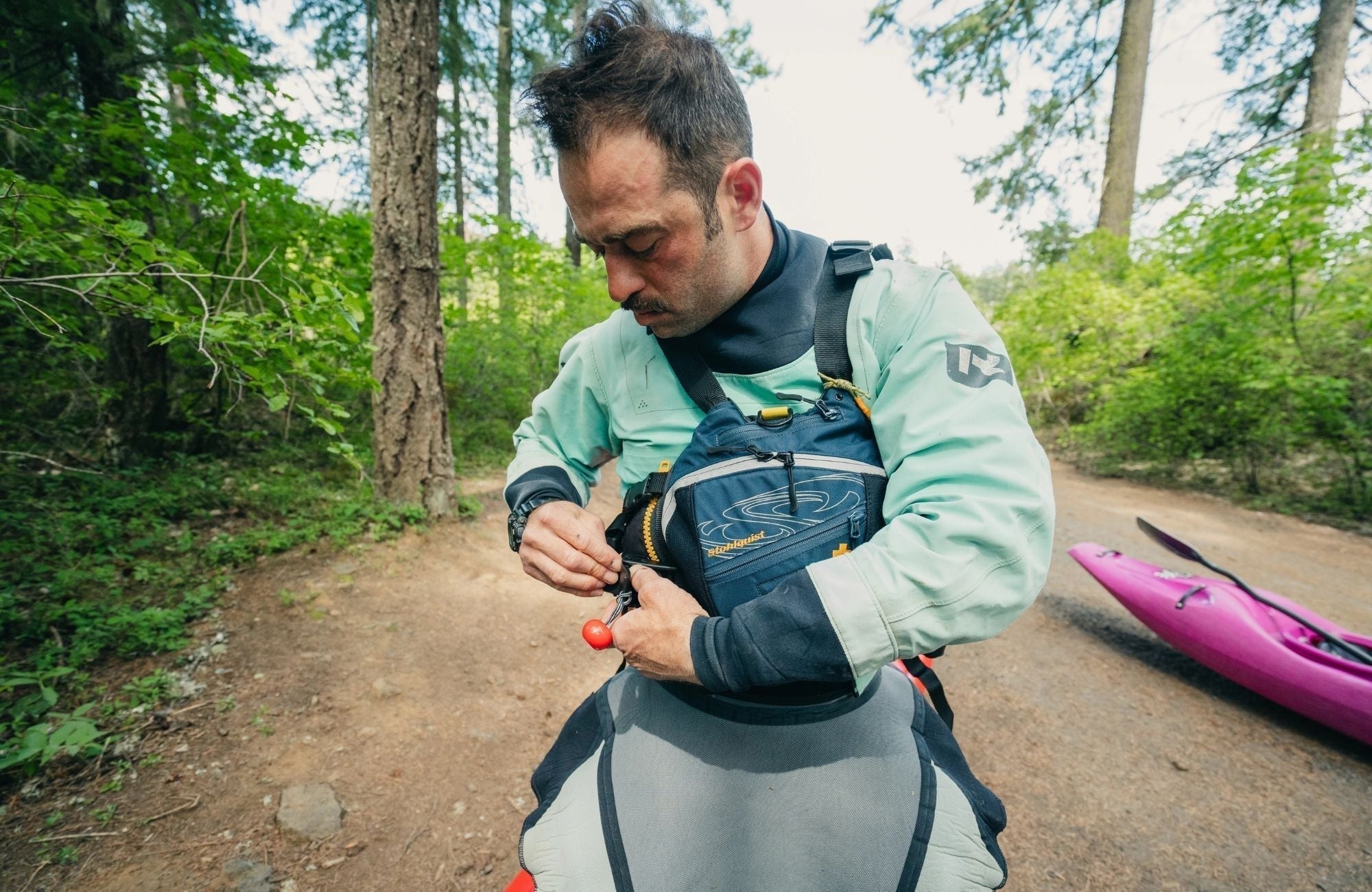 Man wearing Stohlquist kayak life jacket adjusting gear outdoors beside pink kayak