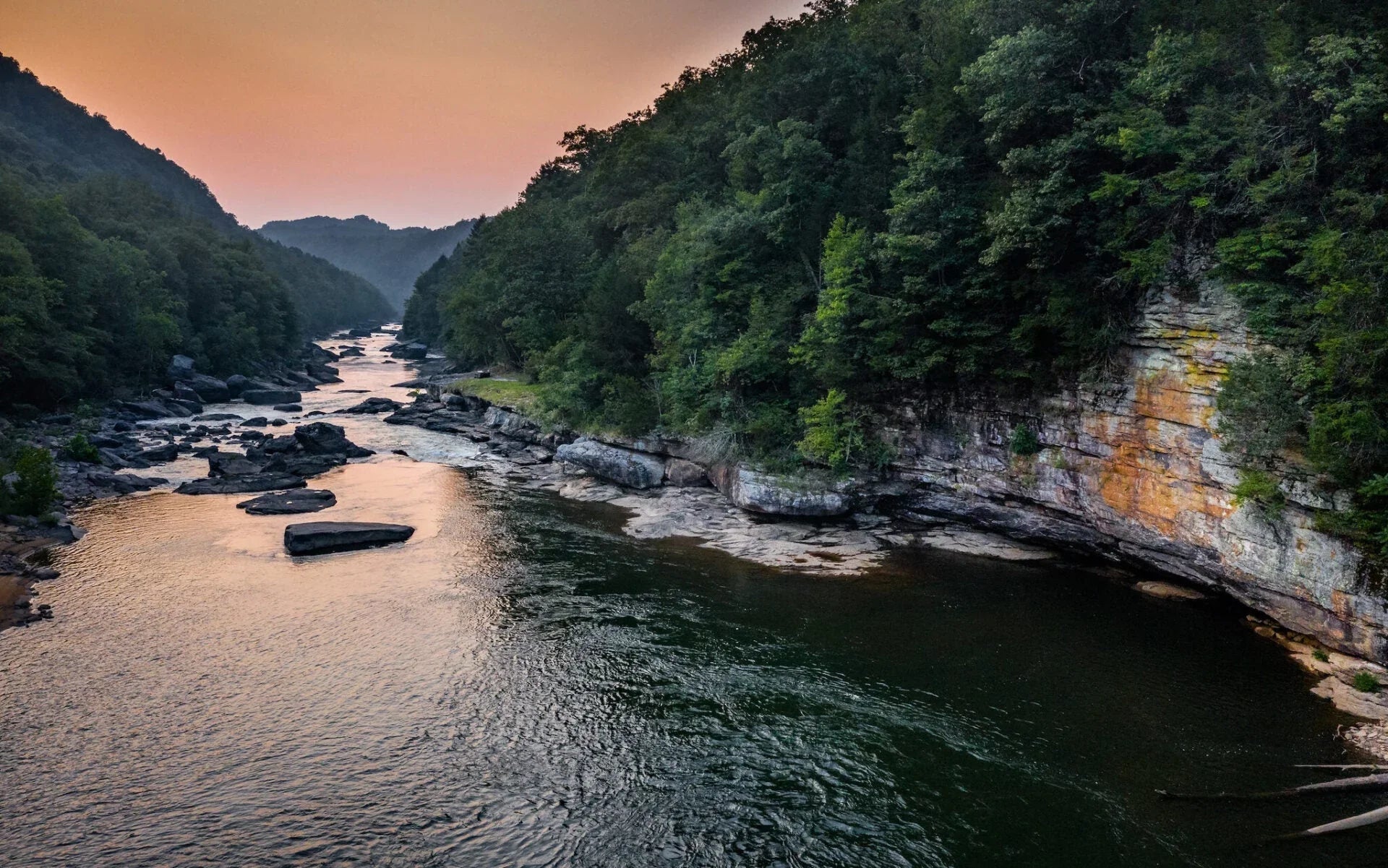 River flowing through forested rocky canyon at sunset with orange sky and green trees