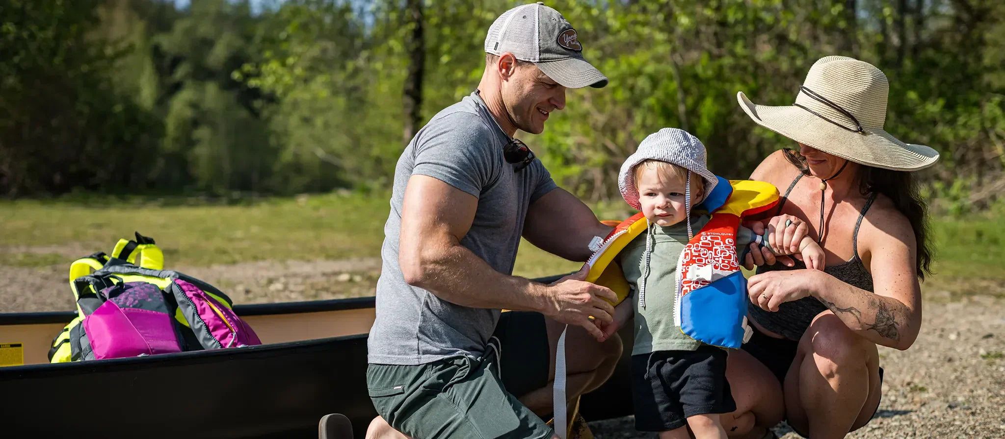 Parents helping toddler wear colorful life jacket near canoe in outdoor nature setting