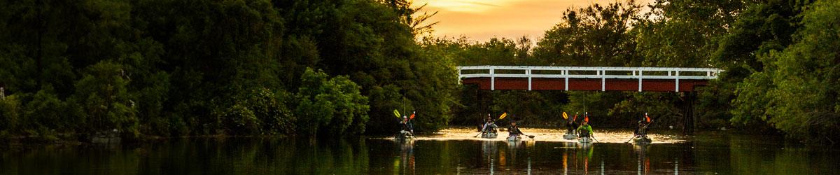 Group kayaking in fishing gear on a calm river under a bridge at sunset, Stohlquist safety jackets