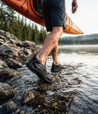 A person walking through shallow water at a rocky lakeshore while wearing black water shoes, with an orange kayak partially visible above and a forested shoreline in the background.